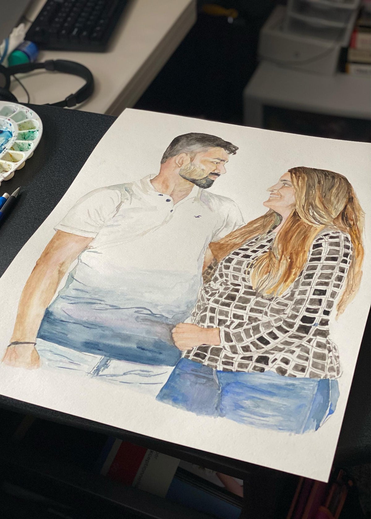 Custom couple portrait being painted in watercolour on a desk, a man and woman embracing beside a paint palette Imagen principal del producto