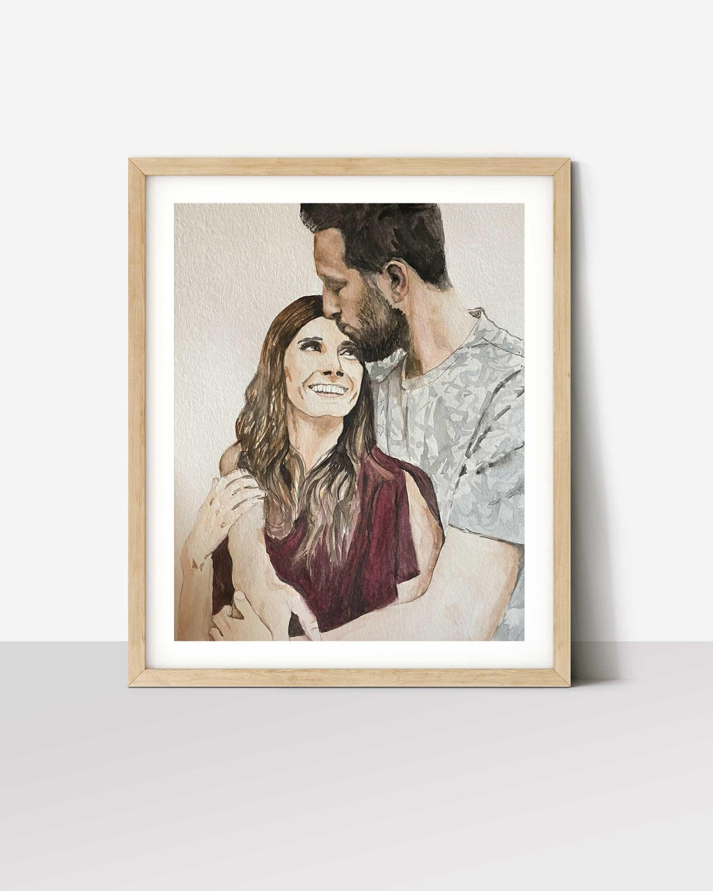Couple portrait painted from a photo, man kissing woman's forehead, in a white-matted oak frame