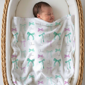 Newborn sleeping under a green and pink bow name blanket in a bassinet